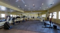 This fitness center is designed to support a full range of workout activities within a single, open-plan floor. The space is generously sized, with high ceilings fitted with recessed lighting and multiple ceiling fans that ensure comfortable air circulation throughout. The walls are finished in a warm yellow tone and lined with large floor-to-ceiling mirrors, which amplify the sense of space and allow users to monitor their form during exercise. The flooring consists of durable rubber or foam tiles, providing cushioning and traction ideal for gym use. The equipment selection includes treadmills, stationary bikes, and an assortment of weight machines and strength training apparatus, catering to both cardio and resistance training needs. Natural light filters in through windows along the perimeter, contributing to an open and energizing atmosphere. The layout is thoughtfully organized, offering clear pathways between machines and ample room for movement. Overall, the fitness center presents a clean, functional, and welcoming environment well-suited for a variety of fitness routines.