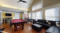 Located at 3530 Brookstone Dr in Cincinnati, Ohio, this single-floor space is designed with entertainment and comfort in mind. The open layout is anchored by dramatic vaulted ceilings that rise to impressive heights in the main gathering area, creating a sense of spaciousness enhanced by large, multi-pane windows dressed with horizontal blinds that allow generous natural light to flood the warm hardwood floors throughout.

The main lounge area features a large sectional leather sofa with matching ottoman, providing ample seating for groups. Adjacent to the lounge, a dedicated game area includes a full-sized pool table and a foosball table, making the space well-suited for social gatherings. A dining nook with a raised pub-style table and chairs sits near glass-panel French doors, and a wall-mounted flat-screen television adds a media component to the area.

The kitchen is outfitted with rich dark espresso cabinetry, granite countertops, a white subway tile backsplash, stainless steel appliances including a refrigerator and microwave, and a double sink — offering full functionality. Crown molding, yellow accent walls, and decorative artwork contribute to the space's inviting, lively ambiance throughout.