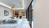 Located at 15635 Memorial Dr in Houston's Memorial neighborhood, this thoughtfully designed single-floor amenity space offers a refined blend of functionality and contemporary style. Light hardwood flooring flows seamlessly throughout, unifying the hallway, dining rooms, and kitchen into a cohesive, welcoming environment.

The open lounge area greets visitors with a striking dome pendant light featuring a bold gold interior, recessed tray ceilings with ambient lighting, and a curated built-in shelving unit with warm accent lighting that houses a workstation and seating nook. A comfortable sectional sofa, accent chairs, and layered rugs contribute to an inviting, collaborative atmosphere.

Two private dining rooms serve as intimate meeting or conferencing spaces, each featuring a dramatic live-edge wood table with industrial steel legs, leather and upholstered seating, and a wall-mounted flat-screen display. Textured blue tile accent walls add visual depth, while floor-to-ceiling black-framed windows flood the rooms with natural light and offer views of the landscaped courtyard outside.

The kitchen area is outfitted with crisp white quartz countertops, soft blue-gray cabinetry with matte black hardware, a gray tile backsplash, and a self-serve beverage station. The space is further enhanced by a wall of black-framed glass panels along the hallway, creating an airy, open-plan feel throughout.