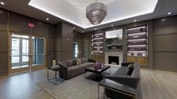 This refined single-floor space exudes contemporary elegance through its carefully curated design and high-end finishes. The lounge area serves as the focal point of the interior, anchored by a classic fireplace framed by a clean mantel, which lends warmth and sophistication to the room. Flanking the fireplace, illuminated built-in shelving units with backlit purple accents provide both display space and a dramatic visual effect, showcasing decorative objects that enhance the overall aesthetic.

Overhead, a layered, tiered metallic chandelier commands attention at the center of the room, casting a warm ambient glow that complements the recessed lighting integrated into the tray ceiling. The ceiling detail itself adds architectural depth and dimension to the space.

The seating arrangement features plush, velvet-toned sofas and chairs in deep charcoal hues, paired with sleek coffee tables that reflect a refined, modern sensibility. A large area rug grounds the conversation area, adding texture and comfort underfoot.

The walls are clad in structured paneling finished in rich, dark tones, creating a cohesive and enveloping atmosphere throughout. Overall, this space presents a polished, upscale environment well-suited for sophisticated social gatherings or relaxed luxury lounging.