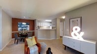 This single-floor residence offers a thoughtfully designed open-concept layout that maximizes functionality and flow. The living area is carpeted and comfortably sized, providing a welcoming space for relaxation and entertaining. A warm accent wall in deep burgundy anchors the dining area, adding a bold visual contrast to the neutral gray and white tones found throughout the rest of the space. The kitchen features a peninsula-style pass-through counter with white upper cabinetry and under-cabinet lighting, making it easy to interact with guests while preparing meals. The clean white cabinetry and bright overhead lighting lend the kitchen a crisp, organized feel. A hallway extends from the main living area, leading to additional rooms, including what appears to be a bathroom with a visible glass-enclosed shower. A textured ceiling adds subtle architectural character throughout. The overall color palette — combining soft grays, warm carpeting, and rich accent tones — creates a balanced and inviting ambiance. The layout is efficient and practical, making excellent use of the available space while maintaining a comfortable, livable atmosphere throughout.