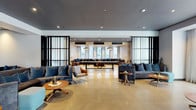 This contemporary amenity space is thoughtfully designed across a single floor, blending sophisticated aesthetics with functional communal living. Polished concrete floors flow seamlessly throughout, unifying the various zones while lending an industrial-chic character to the overall environment.

The main lounge area features plush velvet sofas in deep blue and charcoal tones, arranged around wooden side tables and accented by sleek black-framed glass partitions that define spaces without sacrificing openness. A vibrant teal accent wall anchors a secondary seating area complete with a built-in fireplace, creating a warm and inviting focal point.

A dedicated lounge nook showcases warm wood-paneled ceilings, large floor-to-ceiling windows with sweeping urban views, and a generous wraparound leather sectional sofa adorned with richly colored cushions. Oversized pendant lights hang above, adding a refined finishing touch.

The gourmet kitchen is a standout feature, boasting dark stone countertops, a striking marble-faced island, professional-grade appliances, a herringbone-tiled backsplash, and expansive windows that flood the space with natural light. Ample cabinetry and open shelving ensure both style and practicality.

Together, these areas create a cohesive, design-forward environment ideal for socializing, relaxing, or entertaining.