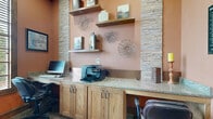 Located in Humble, TX, this business center office is a thoughtfully designed workspace that balances functionality with refined aesthetics. The room features warm terracotta-toned walls complemented by striking stacked stone column accents, creating a professional yet welcoming environment.

A continuous built-in workstation with a smooth granite-style countertop runs along the walls, supported by solid oak cabinetry with ample storage below. The workstation accommodates multiple users simultaneously, equipped with computers and a multifunction printer, making it ideal for everyday business tasks. Ergonomic leather rolling chairs provide comfortable seating throughout.

Floating wooden shelves mounted against the stacked stone backdrop display tasteful decorative pieces, including ceramic vases and sculptural wall art, adding character to the space. A large window fitted with wooden blinds anchors one wall, allowing natural light to filter in while offering a view of the surrounding greenery outdoors. Framed artwork flanks the window, enhancing the room's polished look.

The floor is covered in a neutral, textured carpet that adds warmth underfoot, while the overall layout ensures ease of movement. A doorway connects this space to an adjacent corridor, making it easily accessible. This business center office delivers a complete, professional setting suited for productivity.