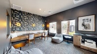 Located at 9600 W Georgia Ave in Glendale, Arizona, this well-appointed single-story amenity space offers a thoughtfully designed environment ideal for both collaborative work and casual relaxation. The layout encompasses six distinct areas that together create a cohesive, modern atmosphere.

The lounge area serves as a central gathering point, featuring bold, graphic-patterned accent wallpaper in dark tones contrasted by warm gold wall sconces. Comfortable seating options — including a built-in banquette with dining tables, open chairs, and oversized bean bag chairs — cater to a variety of uses. Large windows flood the space with natural light while offering views of the surrounding outdoor courtyard, complete with palm trees and landscaped greenery.

A dedicated private conference room provides a more formal setting, centered around a striking live-edge wood table surrounded by upholstered gray chairs. A wall-mounted display screen, textured accent wall panels, and a curated gallery of large-scale framed artwork lend the room a professional yet creative character. Recessed lighting and carpeted flooring maintain a polished, focused environment.

Additional areas within the space include workstations and support rooms, all finished with consistent neutral gray tones, recessed lighting, and carpet flooring. Throughout, the design balances contemporary aesthetics with practical functionality, making this a versatile and inviting community facility.
