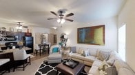 This single-floor residence offers a thoughtfully designed open-concept layout that seamlessly connects the living and dining areas, creating an inviting and functional living environment. The living area is anchored by a comfortable sectional sofa, a tufted ottoman coffee table, and a patterned area rug, all set beneath a ceiling fan with integrated lighting. A large, colorful abstract artwork adorns the wall, adding a bold visual focal point to the space. The room is finished with neutral-toned walls and light-colored flooring that enhances the sense of openness and brightness throughout. An accent chair with a coordinating patterned design adds an additional seating option and a touch of personality. The dining area is situated adjacent to the living space, featuring a dark bistro-style table with matching bar-height chairs, creating an intimate setting for meals. Decorative elements including mirrored accents, vases, and curated accessories are thoughtfully placed throughout, contributing to a cohesive and polished aesthetic. The overall ambiance of the space is modern and welcoming, with a neutral color palette accented by teal and earthy tones that lend warmth and character to every corner.