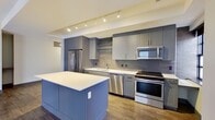This thoughtfully designed 2-bedroom, 1-bathroom residence offers a seamless blend of modern aesthetics and functional living across a single floor. The open-concept kitchen and living area serves as the heart of the home, featuring rich dark hardwood-style flooring that extends throughout. The kitchen is outfitted with sleek grey cabinetry, a subway tile backsplash, stainless steel appliances including a French door refrigerator, gas range, built-in microwave, and dishwasher, all anchored by a central island with a crisp white countertop. Track lighting overhead adds a contemporary touch. The spacious living room is bathed in natural light through multiple large windows with dark frames, lending an urban character to the open layout. A wide hallway with recessed lighting connects the living spaces to the private quarters. Both bedrooms feature plush carpeting and ceiling fans, with the primary bedroom offering direct access to the full bathroom and a walk-in closet. The bathroom is finished with white subway tile in the glass-enclosed shower, tile flooring, and a vanity with dark cabinetry. A second closet with built-in shelving provides additional storage. The space presents a clean, contemporary feel throughout with a cohesive neutral color palette and dark trim accents.