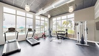 Located at 2201 N Australian Ave in West Palm Beach, FL, this fitness center offers a well-equipped, light-filled workout environment on a single floor. Large, floor-to-ceiling windows line multiple walls, flooding the space with natural light and providing sweeping views of a resort-style pool, palm trees, and open sky — creating an energizing atmosphere for exercise. The interior features a durable dark rubber flooring suited for high-traffic use, and an exposed industrial-style ceiling with visible ductwork and grid paneling adds an urban, modern aesthetic. Pendant lighting provides additional illumination throughout. The facility is equipped with a variety of cardio machines, including treadmills and a spin bike, as well as strength training equipment such as a cable machine, dumbbells with a multi-tier rack, an elliptical, and a weight bench. A large flat-screen TV and a full-length mirror are also present. One wall is adorned with bold motivational typography — featuring words like "Achieve," "Endurance," "Confident," and "Determination" — adding an inspiring visual element to the space. A direct exterior door provides easy access to the surrounding outdoor amenities, seamlessly connecting indoor fitness with the outdoor environment.