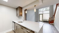 Located at 1 Shearwood Place in New Rochelle, NY, this thoughtfully designed two-story residence offers a comfortable and modern living experience. The main level provides a generous open-concept layout, where the kitchen seamlessly flows into the living area. The kitchen is outfitted with warm wood-toned cabinetry, a sleek white quartz countertop with a waterfall island, a stainless steel sink, and stylish pendant lighting that adds a contemporary touch. Light hardwood-style flooring runs throughout, complementing the neutral gray walls and bright, airy atmosphere. A large window on the main level allows natural light to fill the space, while a sliding glass door offers outdoor views and easy access. A classic wood-banister staircase leads to the upper level, where the bedroom is situated. The bedroom is a comfortable size, featuring tall ceilings, large windows with vertical blinds that frame greenery views, and continued hardwood-style flooring. The full bathroom is elegantly appointed with a wood-toned vanity, white quartz countertop, and a tub-shower combination adorned with stunning marble-look tile surround and brushed gold fixtures. A dedicated closet and hallway complete the upper floor, ensuring practical storage and flow throughout this well-appointed home.