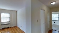 This single-floor space presents a straightforward and functional layout characterized by its clean, minimalist aesthetic. The interior walls are finished in a crisp, neutral white tone that lends a bright and open feel to the space, while the warm-toned hardwood flooring runs throughout, adding a natural warmth that contrasts pleasantly with the light walls. White baseboards frame the perimeter of the rooms, providing a polished, cohesive look. A white-trimmed interior door is visible along one wall, offering access to an adjoining room or closet and contributing to the space's tidy, well-maintained appearance. A light switch plate is positioned conveniently along the wall, and subtle electrical outlets are recessed near the baseboard. The ceiling height appears generous, contributing to an airy atmosphere throughout. The overall ambiance of the space is clean and move-in ready, with a neutral palette that provides a versatile foundation for a variety of furnishing styles and personal touches.