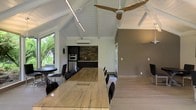 This well-appointed single-floor space makes an immediate impression with its dramatic vaulted ceiling, finished in crisp white-painted wood beams and rafters that lend an airy, open quality to the interior. A large ceiling fan anchors the main living and dining area, complementing the natural light that streams in through generous windows framing lush tropical greenery outside.

The centerpiece of the main area is a substantial wood dining table with seating for a large group, offering an ideal setting for gatherings. Sleek black chairs provide a contemporary contrast to the warm, natural tones of the hardwood-style flooring that runs throughout the space.

The kitchen features dark cabinetry paired with a bold stacked-stone backsplash, creating a striking focal point. Built-in appliances, including a wall oven and refrigerator, are seamlessly integrated into the cabinetry for a polished, functional layout. A wall-mounted split air conditioning unit ensures year-round comfort.

A smaller bistro-style table near the window offers an intimate alternative seating nook bathed in natural light. The overall ambiance balances rustic architectural charm with modern finishes, resulting in a space that feels both inviting and refined.