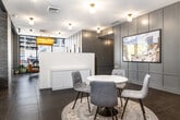 This thoughtfully designed single-floor office suite offers a refined blend of contemporary and classic aesthetics across three offices and a connecting hallway. The main reception area immediately impresses with its open layout, anchored by a sleek white reception desk and a round marble-top table surrounded by upholstered chairs set atop a patterned area rug — ideal for welcoming guests or informal meetings. Large floor-to-ceiling windows along the exterior walls flood the space with natural light while offering clear views to the street, creating an energizing and transparent atmosphere. Dark porcelain tile flooring runs throughout, providing a sophisticated foundation that contrasts elegantly with the white walls and gray wainscoted paneling. Warm brass globe light fixtures are mounted across the ceiling, lending a polished, boutique character to the space. A dedicated mailroom hallway features a built-in row of gold mailboxes, a prominent wall clock, and an open shelving unit, offering practical functionality. Additional office areas are defined by rich wooden double doors with glass panels, open industrial-style shelving, and curated décor including framed artwork and indoor greenery. The overall ambiance is professional yet welcoming, combining clean modern lines with warm, character-rich details throughout.