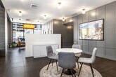This thoughtfully designed single-floor office suite offers a refined blend of contemporary and classic aesthetics across three offices and a connecting hallway. The main reception area immediately impresses with its open layout, anchored by a sleek white reception desk and a round marble-top table surrounded by upholstered chairs set atop a patterned area rug — ideal for welcoming guests or informal meetings. Large floor-to-ceiling windows along the exterior walls flood the space with natural light while offering clear views to the street, creating an energizing and transparent atmosphere. Dark porcelain tile flooring runs throughout, providing a sophisticated foundation that contrasts elegantly with the white walls and gray wainscoted paneling. Warm brass globe light fixtures are mounted across the ceiling, lending a polished, boutique character to the space. A dedicated mailroom hallway features a built-in row of gold mailboxes, a prominent wall clock, and an open shelving unit, offering practical functionality. Additional office areas are defined by rich wooden double doors with glass panels, open industrial-style shelving, and curated décor including framed artwork and indoor greenery. The overall ambiance is professional yet welcoming, combining clean modern lines with warm, character-rich details throughout.