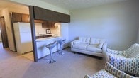 This single-floor space is designed with an open and functional layout that seamlessly connects the kitchen and living area. The kitchen is framed by dark wood cabinetry overhead, providing a warm contrast to the light-colored countertops and white appliances visible within. A pass-through counter extends into the living area, creating a casual bar-style seating space accommodated by two modern metallic barstools, making it ideal for quick dining or entertaining.

The living area features wall-to-wall carpeting in a neutral tone, contributing to a cozy and comfortable atmosphere. A light-colored loveseat and additional seating offer a relaxed arrangement suited for everyday living. The walls are painted in a soft, muted green, giving the space a calm and understated feel. A textured ceiling adds a subtle architectural detail throughout.

The pass-through design between the kitchen and living room enhances the sense of openness, allowing natural interaction between the two spaces while maintaining a degree of separation. Overall, the layout is practical and efficiently organized, making good use of the available space.