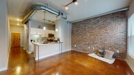 This single-floor unit at MAA Piedmont Park showcases an urban loft aesthetic defined by a striking combination of industrial and contemporary design elements. Polished stained concrete floors flow continuously throughout, reflecting light and adding warmth to the open layout. Exposed silver ductwork runs prominently across the ceilings, reinforcing the industrial character of the space, while track lighting provides functional illumination throughout.

The open living area is anchored by a full-height exposed brick accent wall, lending texture and visual depth to the room. Natural light enters through a window at the far end of the space, brightening the area. The kitchen is thoughtfully designed with white cabinetry, granite countertops, a subway tile backsplash, and stainless steel appliances including a microwave, range, and refrigerator. A peninsula with a metal-topped counter serves as both a workspace and casual dining area, with pendant lighting overhead.

A hallway connects the living and kitchen areas to the bathroom and additional rooms. The bathroom features a tub-shower combination, white vanity cabinetry with granite countertops, a large mirror, and continued polished concrete flooring, maintaining design consistency throughout the unit.