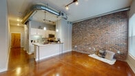 This single-floor unit at MAA Piedmont Park showcases an urban loft aesthetic defined by a striking combination of industrial and contemporary design elements. Polished stained concrete floors flow continuously throughout, reflecting light and adding warmth to the open layout. Exposed silver ductwork runs prominently across the ceilings, reinforcing the industrial character of the space, while track lighting provides functional illumination throughout.

The open living area is anchored by a full-height exposed brick accent wall, lending texture and visual depth to the room. Natural light enters through a window at the far end of the space, brightening the area. The kitchen is thoughtfully designed with white cabinetry, granite countertops, a subway tile backsplash, and stainless steel appliances including a microwave, range, and refrigerator. A peninsula with a metal-topped counter serves as both a workspace and casual dining area, with pendant lighting overhead.

A hallway connects the living and kitchen areas to the bathroom and additional rooms. The bathroom features a tub-shower combination, white vanity cabinetry with granite countertops, a large mirror, and continued polished concrete flooring, maintaining design consistency throughout the unit.