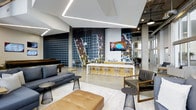 This vibrant clubhouse is a thoughtfully designed social hub spread across a single floor, blending contemporary aesthetics with welcoming functionality. The space is anchored by a dramatic large-scale mural spanning an entire wall, rendered in bold dark tones with graphic map-like patterns that serve as a striking artistic focal point. Adjacent to the mural is a sleek bar area finished in warm gold tones, lined with modern wire bar stools that invite casual conversation and gathering. The open-plan layout is illuminated by recessed linear LED lighting in the ceiling, which casts a clean, modern glow throughout the space. Exposed concrete columns add an industrial character that contrasts beautifully with the polished finishes elsewhere. The lounge area is furnished with a rich leather ottoman, charcoal sofas, and accent chairs paired with wire-frame side tables, creating a comfortable and stylish setting for socializing. A wall-mounted flat-screen television enhances the entertainment experience. Toward the rear, a pool or game table adds a recreational dimension to the space. Large glass doors or windows allow natural light to filter in, connecting the interior to the surrounding environment. The overall ambiance is urban, energetic, and inviting — a space designed to bring people together.