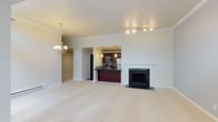 This single-floor residence offers a thoughtfully designed open-concept layout that seamlessly connects the main living areas. The spacious living room is anchored by a classic fireplace framed by a crisp white mantel and dark tile surround, adding a refined focal point to the space. Soft, neutral-toned carpeting runs throughout, complementing the clean white crown molding and baseboards that lend a polished, cohesive feel.

A decorative chandelier with warm lighting defines the dining area, adding an elegant touch to everyday gatherings. The kitchen is positioned adjacent to the living space and features rich dark wood cabinetry, a mosaic tile backsplash, stainless steel appliances — including a refrigerator and microwave — and a dark granite-topped breakfast bar that provides both workspace and casual seating. Track lighting overhead ensures the kitchen is well-lit and functional.

A hallway visible from the main living area leads further into the residence, hinting at additional private spaces beyond. The overall ambiance is warm and inviting, characterized by neutral tones, quality finishes, and a layout designed for comfortable everyday living.