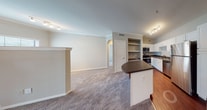 This single-floor residence offers a well-designed layout with distinct living zones connected by a cohesive neutral color palette and a mix of hardwood-style and carpeted flooring. The kitchen is a functional highlight, featuring white cabinetry, dark granite-style countertops, and a full suite of stainless steel appliances including a refrigerator, electric range, microwave, and dishwasher. A peninsula with a curved countertop extends into the adjacent space, providing additional prep area and casual seating. A built-in shelving nook adjacent to the kitchen offers convenient storage or workspace potential. The living area is generously sized, anchored by a white-framed fireplace and a sliding glass door that allows natural light to filter in through vertical blinds. Crown molding and a ceiling fan add architectural detail to this space. The bedroom is carpeted and bright, benefiting from a large window with outdoor views and a ceiling fan overhead. The bathroom includes a vanity with white cabinetry, dark countertops, a large mirror with overhead lighting, and a tub-shower combination with glass enclosure. Overall, the space presents a practical and comfortable layout with consistent finishes throughout.