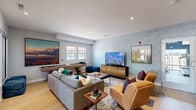This well-appointed single-floor community space is thoughtfully designed to serve as both a welcoming lounge and a functional leasing center. The layout flows seamlessly across two primary areas, each offering a distinct purpose while maintaining a cohesive modern aesthetic throughout.

The living room is a standout gathering space, anchored by a geometric patterned accent wall in soft blue-grey tones that adds visual depth and personality. Light hardwood flooring runs throughout, complementing the neutral palette. Comfortable seating, including plush sofas and cognac leather chairs, is arranged around dual flat-screen displays, while a shuffleboard table adds a recreational element. Recessed lighting and natural light from multiple windows brighten the space.

The dining area is equally inviting, featuring a round wooden dining table surrounded by upholstered chairs beneath a sculptural multi-arm chandelier. A sliding glass door opens to an outdoor view, filling the room with natural light. An adjacent kitchenette with bold navy cabinetry and pendant lighting provides a practical refreshment station.

The reception area is defined by a fluted-panel desk, warm leather guest chairs, and a striking overhead fixture, with glass partitions offering open sightlines into the lounge. Together, these spaces create an engaging, community-focused environment with a polished yet approachable character.