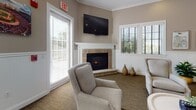 Located at 4980 N Main St in Fall River, Massachusetts, this single-story community clubhouse offers a well-appointed gathering space designed to foster connection and comfort. The interior is finished with warm neutral tones, crown molding, and classic wainscoting throughout, creating a cohesive and inviting aesthetic.

The main lounge area serves as the heart of the space, featuring a handsome corner fireplace with a stone surround and white mantel, wall-mounted television, and plush upholstered armchairs arranged for easy conversation. Multiple windows dressed with plantation shutters allow generous natural light to fill the room.

Adjacent to the lounge, a dedicated dining and meeting area provides a comfortable setting for group gatherings, complete with a dining table, coordinating chairs, and additional natural light from surrounding windows. A billiards table anchors the central open area, offering a recreational focal point for residents.

The fully equipped kitchen features white cabinetry, granite-toned countertops, stainless steel appliances including a full-size refrigerator and dishwasher, tile flooring, and an extended bar-height counter with seating — ideal for entertaining. An entryway corridor, accented by a vibrant cityscape mural of Fall River, connects the various zones and adds a sense of local character to the overall design.