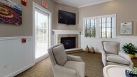 Located at 4980 N Main St in Fall River, Massachusetts, this single-story community clubhouse offers a well-appointed gathering space designed to foster connection and comfort. The interior is finished with warm neutral tones, crown molding, and classic wainscoting throughout, creating a cohesive and inviting aesthetic.

The main lounge area serves as the heart of the space, featuring a handsome corner fireplace with a stone surround and white mantel, wall-mounted television, and plush upholstered armchairs arranged for easy conversation. Multiple windows dressed with plantation shutters allow generous natural light to fill the room.

Adjacent to the lounge, a dedicated dining and meeting area provides a comfortable setting for group gatherings, complete with a dining table, coordinating chairs, and additional natural light from surrounding windows. A billiards table anchors the central open area, offering a recreational focal point for residents.

The fully equipped kitchen features white cabinetry, granite-toned countertops, stainless steel appliances including a full-size refrigerator and dishwasher, tile flooring, and an extended bar-height counter with seating — ideal for entertaining. An entryway corridor, accented by a vibrant cityscape mural of Fall River, connects the various zones and adds a sense of local character to the overall design.
