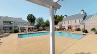 This outdoor pool area offers a well-appointed retreat designed for relaxation and recreation. The centerpiece is a generously sized swimming pool with clear blue water, accessible via built-in entry steps and metal handrails. The surrounding deck is finished in a textured, aggregate concrete surface that provides both durability and a warm, neutral aesthetic underfoot.

Ample deck space accommodates multiple rows of sling-style chaise lounges, ideal for sunbathing throughout the day. White pergola structures are positioned poolside, adding architectural interest while providing shaded areas furnished with additional seating and red market umbrellas for sun protection. Hanging planters with trailing greenery accent the pergolas, adding a touch of natural detail.

The pool area is enclosed by a black wrought iron fence, ensuring security while maintaining open sightlines to the surrounding landscaping. Mature trees and flowering shrubs frame the perimeter, offering natural shade and a lush backdrop. A brick building with green shutters and French doors is visible adjacent to the pool, providing convenient access to interior amenities. The overall atmosphere is clean, open, and well-maintained under clear skies.