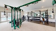 Located at 101 Lemon Dr in Lake Saint Louis, MO, this fitness center offers a generously sized, single-floor workout facility designed to support a full spectrum of exercise routines. The space is thoughtfully divided into distinct functional zones, providing an organized and efficient layout for users of all fitness levels.

The cardio area lines one side of the room and is outfitted with multiple treadmills, elliptical trainers, a rowing machine, and a balance trainer. Large windows along this section flood the space with natural light and offer pleasant outdoor views of the surrounding landscape, while wall-mounted televisions keep users entertained during workouts.

The strength training zone is equally impressive, anchored by a prominent Life Fitness multi-station cable machine in a distinctive green finish. A dedicated free weights area features a full dumbbell rack with an adjacent large mirror wall, enhancing the sense of depth in the room. Power racks with Olympic benches and weight plates provide ample options for compound lifts.

The flooring transitions between light carpet in the cardio zone and durable rubber flooring in the strength area, clearly delineating each section. Recessed lighting throughout ensures consistent illumination across the entire space. Stylish patterned accent wallpaper adds a modern design touch, while glass doors open to an outdoor pool area, connecting the indoor fitness experience to exterior amenities.