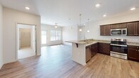 This single-story residence at 1585 E Cornelius Harnett Blvd in Lillington, NC offers a thoughtfully designed open-concept layout with a neutral, modern aesthetic throughout. The kitchen serves as a central hub of the home, featuring rich dark cabinetry, granite countertops, stainless steel appliances, and a peninsula with bar seating illuminated by pendant lighting overhead. Recessed lighting and an open sightline connect the kitchen seamlessly to the main living area, which is finished with light wood-style flooring, a ceiling fan, and a glass door providing access to the outdoors and inviting natural light inside.

The bedrooms are carpeted for comfort and equipped with ceiling fans. Multiple windows with blinds allow generous natural light and outdoor views, creating a bright and airy atmosphere. At least one bedroom benefits from a corner window configuration, further enhancing the sense of openness.

The bathroom is well-appointed with dark wood cabinetry, granite countertops, a large mirror, and matching light wood-style flooring. A full tub and shower combo with a brushed nickel curtain rod completes the bath. An in-unit laundry area is also visible, adding everyday convenience to this clean, move-in-ready home.