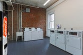 Located at 104 W 17th St in Chattanooga, Tennessee, this single-floor laundry facility combines industrial character with practical functionality. The space is defined by its striking exposed brick walls, which lend a raw, historic aesthetic that contrasts with the clean white painted surfaces throughout the room.

The flooring consists of dark gray tile laid in a grid pattern, providing a durable and easy-to-maintain surface suited to the facility's purpose. Multiple commercial-grade washing machines and dryers are arranged along the walls, offering ample laundry capacity. A utility sink is also present, adding to the room's functional layout. Overhead, exposed piping and conduit run along the walls and ceiling, reinforcing the space's utilitarian character.

Natural light enters through double-hung windows, brightening the interior and offering views of the surrounding neighborhood. A Coca-Cola vending machine adds a practical amenity near one of the entry doors. The space features multiple entry points, including a wood-paneled interior door and a standard paneled door marked for employee access. Fire safety equipment is wall-mounted and clearly visible. Overall, the space presents a no-frills, well-equipped shared laundry facility with notable historic architectural details.