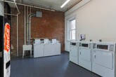 Located at 104 W 17th St in Chattanooga, Tennessee, this single-floor laundry facility combines industrial character with practical functionality. The space is defined by its striking exposed brick walls, which lend a raw, historic aesthetic that contrasts with the clean white painted surfaces throughout the room.

The flooring consists of dark gray tile laid in a grid pattern, providing a durable and easy-to-maintain surface suited to the facility's purpose. Multiple commercial-grade washing machines and dryers are arranged along the walls, offering ample laundry capacity. A utility sink is also present, adding to the room's functional layout. Overhead, exposed piping and conduit run along the walls and ceiling, reinforcing the space's utilitarian character.

Natural light enters through double-hung windows, brightening the interior and offering views of the surrounding neighborhood. A Coca-Cola vending machine adds a practical amenity near one of the entry doors. The space features multiple entry points, including a wood-paneled interior door and a standard paneled door marked for employee access. Fire safety equipment is wall-mounted and clearly visible. Overall, the space presents a no-frills, well-equipped shared laundry facility with notable historic architectural details.