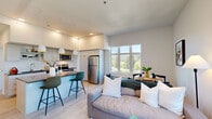 This well-appointed 2-bedroom, 2-bathroom residence offers a thoughtfully designed single-floor layout with a clean, contemporary aesthetic. Light-toned hardwood-style flooring and crisp white walls run throughout, creating a bright and cohesive atmosphere.

The open-concept kitchen and living area serves as the heart of the home. The kitchen features flat-front white cabinetry, dark countertops, a white tile backsplash, stainless steel appliances, and a breakfast bar with pendant lighting. The living area is generously sized with large windows that invite abundant natural light and offer pleasant treetop and neighborhood views. A dedicated dining space sits adjacent, completing the open layout.

Both bedrooms are well-proportioned, featuring large multi-pane windows that frame outdoor greenery and surrounding scenery. The primary bedroom offers a notably spacious footprint, while the second bedroom provides a comfortable and equally light-filled space.

The two bathrooms are finished in a modern style, with one featuring a glass-enclosed shower, white subway tile surround, a wide vanity with dark countertop, and ample cabinetry. The unit also includes a dedicated laundry room with a stacked washer and dryer. A welcoming entry hallway with recessed lighting completes this functional and stylish residence.