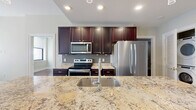 This thoughtfully designed 2-bedroom, 2-bathroom residence offers a functional and stylish single-floor layout. The entrance opens to a flowing open-concept living and kitchen area, where rich hardwood floors extend throughout the main living spaces. The kitchen serves as a central hub, anchored by a large island with striking granite countertops, an undermount sink with a modern faucet, and elegant pendant lighting overhead. Dark espresso cabinetry reaches toward the ceiling, complemented by a tile backsplash and a full suite of stainless steel appliances. The living area benefits from natural light streaming through large windows with views of surrounding architecture. Both bedrooms feature the same warm hardwood flooring, with the secondary bedroom offering dual windows. Organized closet systems with built-in shelving and hanging rods are found in each bedroom. The primary bathroom is finished with marble-style tile flooring and walls, a floating navy vanity with dual sinks, gold hardware accents, and a glass-enclosed shower. The second bathroom echoes the marble aesthetic with a tub-shower combination. An in-unit laundry area adds everyday convenience to this well-rounded residence.