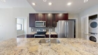 This thoughtfully designed 2-bedroom, 2-bathroom residence offers a functional and stylish single-floor layout. The entrance opens to a flowing open-concept living and kitchen area, where rich hardwood floors extend throughout the main living spaces. The kitchen serves as a central hub, anchored by a large island with striking granite countertops, an undermount sink with a modern faucet, and elegant pendant lighting overhead. Dark espresso cabinetry reaches toward the ceiling, complemented by a tile backsplash and a full suite of stainless steel appliances. The living area benefits from natural light streaming through large windows with views of surrounding architecture. Both bedrooms feature the same warm hardwood flooring, with the secondary bedroom offering dual windows. Organized closet systems with built-in shelving and hanging rods are found in each bedroom. The primary bathroom is finished with marble-style tile flooring and walls, a floating navy vanity with dual sinks, gold hardware accents, and a glass-enclosed shower. The second bathroom echoes the marble aesthetic with a tub-shower combination. An in-unit laundry area adds everyday convenience to this well-rounded residence.