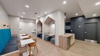 Located at 4125 Chestnut St in Philadelphia's University City neighborhood, this single-floor communal space is thoughtfully designed to support both focused work and casual gathering. The interior features light wood-tone flooring throughout, creating a warm and cohesive aesthetic that complements the clean white walls and dark navy accent panels.

A standout feature of the space is its pair of arch-shaped private nook seating areas, upholstered in soft teal and gray tones, providing semi-enclosed spots ideal for quiet study or small group conversations. Along one wall, a vibrant blue tufted banquette runs the length of the room, accompanied by white-topped tables and natural wood chairs for comfortable dining or group work.

A central island-style workstation with a light wood finish and built-in storage offers bar-height seating along one side, positioned adjacent to a bold, colorful mural that adds an energetic, urban artistic element to the space. A large mounted flat-screen television is integrated into a dark accent column, offering an entertainment focal point within the lounge area.

Recessed lighting throughout ensures the space is well-lit, while motivational wall signage and curated artwork contribute to an upbeat, community-oriented atmosphere. The layout flows openly, accommodating a variety of uses from individual study to social interaction.