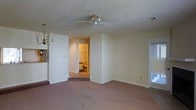 This single-floor residence presents a straightforward and functional layout centered around a spacious main living area finished with wall-to-wall carpeting in a neutral tone. The room is illuminated by a combination of a ceiling fan with an integrated light fixture and an elegant brass chandelier, providing versatile lighting options. Smooth white walls and standard-height ceilings contribute to a clean, understated aesthetic throughout.

A doorway visible from the main living area leads into what appears to be a bathroom or en-suite, warmly lit and featuring visible fixtures including what appears to be a toilet and shelving, suggesting practical storage. A partial view to the left hints at an adjacent area, possibly a kitchenette or utility space, indicated by upper cabinetry.

Natural light filters into the space through a window fitted with vertical blinds on the right side of the room, offering a connection to the outdoors. The overall color palette — soft whites, warm beiges, and muted carpeting — creates a calm, neutral ambiance throughout the residence. The layout appears efficient and well-suited for comfortable single-floor living.