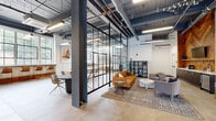 Located at 2501 Oakford Street in Philadelphia, this single-floor commercial space masterfully combines industrial architecture with contemporary design. The open floor plan features soaring ceilings with exposed ductwork and dark-painted structural elements, creating a raw yet polished aesthetic throughout.

The main area is anchored by a comfortable lounge with a warm leather sofa, upholstered seating, and layered rugs atop large-format tile flooring. A striking floor-to-ceiling steel-framed glass partition wall separates the lounge from an adjacent conference or meeting area, maintaining visual connectivity while offering acoustic separation. Natural light floods the space through expansive black-framed industrial windows, providing neighborhood views.

A standout feature is the herringbone wood accent wall paired with a built-in kitchenette area, complete with counter seating and a beverage station. Warm wood stairs with a sleek metal railing provide access to additional levels. A separate entry vestibule with large glass sliding doors and casual seating offers a welcoming transition into the space.

Throughout, the design balances reclaimed wood details, clean white walls, modern lighting, and open sightlines, resulting in a functional, stylish, and highly adaptable commercial environment.
