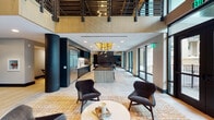 Located at 3700 National Dr in Raleigh, NC, this two-floor amenity space delivers a compelling blend of modern design and versatile functionality. The main level opens into a dramatic double-height lounge anchored by floor-to-ceiling black-framed windows that flood the interior with natural light and offer views of an outdoor terrace and pool area. Bold cylindrical columns in matte black punctuate the open-plan layout, complementing a sleek full kitchen outfitted with dark cabinetry, marble backsplash, and a large island beneath statement pendant lighting. Curated seating areas with designer lounge chairs invite relaxed gathering throughout. A dedicated coworking zone features long shared worktables alongside glass-partitioned private rooms, catering to a range of work styles. Breakout nooks positioned at corner windows provide intimate settings with views of the surrounding courtyard. The lower level transitions to an industrial aesthetic with exposed black ceilings, concrete floors, and a versatile maker studio equipped with a long work bench, cabinetry, and a communal table. A fitness area with cardio equipment and an adjoining conference room round out the offerings. Curated artwork displayed throughout adds a cultural dimension to this well-appointed, multi-use amenity environment.