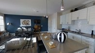 This single-floor space showcases an open-concept layout that connects the kitchen, dining, and living areas into one cohesive, functional environment. The kitchen is appointed with white shaker-style cabinetry, sleek chrome hardware, a classic white subway tile backsplash, and dark countertops that add a sophisticated contrast. Pendant lights hang above the extended kitchen island, which doubles as a dining surface with seating for multiple guests, making it ideal for both casual meals and entertaining.

The dining area flows naturally from the kitchen, featuring a generously sized island countertop dressed with decorative wooden serving boards and a striking black-and-white ceramic vase as a focal point. The living area adjacent to the dining space is anchored by a bold deep-blue accent wall, adding depth and personality to the room. A patterned area rug, coordinating sofa, and curated wall art contribute to the cohesive aesthetic. Wood-look flooring runs throughout, tying the spaces together, while recessed lighting and pendant fixtures provide layered illumination across the entire open floor plan.