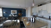 This single-floor space showcases an open-concept layout that connects the kitchen, dining, and living areas into one cohesive, functional environment. The kitchen is appointed with white shaker-style cabinetry, sleek chrome hardware, a classic white subway tile backsplash, and dark countertops that add a sophisticated contrast. Pendant lights hang above the extended kitchen island, which doubles as a dining surface with seating for multiple guests, making it ideal for both casual meals and entertaining.

The dining area flows naturally from the kitchen, featuring a generously sized island countertop dressed with decorative wooden serving boards and a striking black-and-white ceramic vase as a focal point. The living area adjacent to the dining space is anchored by a bold deep-blue accent wall, adding depth and personality to the room. A patterned area rug, coordinating sofa, and curated wall art contribute to the cohesive aesthetic. Wood-look flooring runs throughout, tying the spaces together, while recessed lighting and pendant fixtures provide layered illumination across the entire open floor plan.