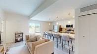 This well-appointed single-floor unit offers a thoughtfully designed open-concept layout that seamlessly connects the living and kitchen areas. The kitchen is a standout feature, boasting white upper cabinetry, warm wood-toned lower cabinets, stainless steel appliances including a refrigerator, microwave, and range, and a generous island with a white quartz countertop and seating for multiple bar-height chairs. Pendant lighting above the island adds a refined touch, while recessed lighting illuminates the entire space. Light hardwood-style flooring runs throughout the main living areas, complementing the neutral, bright palette. The living area features soaring ceilings with a raised detail and a large window that brings in abundant natural light and outdoor views. A cozy seating area provides a comfortable space to relax. The unit includes two carpeted bedrooms, each offering a serene, minimalist aesthetic with ample space. A full bathroom features a combination tub and shower, a wood-tone vanity with a marble-style countertop, and light flooring. A frosted-glass interior door and a bold red entry door add distinctive character to the hallway. In-unit laundry adds everyday convenience to this modern, move-in-ready space.