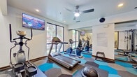 This fitness center is a thoughtfully designed workout facility that offers a comprehensive range of equipment to support diverse exercise routines. The space is finished with a striking hexagonal-patterned carpet in shades of blue, teal, and grey, lending a modern and energetic aesthetic throughout.

The cardio zone features treadmills, a stationary bike, and an elliptical machine, all positioned near large floor-to-ceiling windows that flood the room with natural light and provide views of the surrounding greenery. A ceiling fan overhead ensures comfortable air circulation during workouts.

The strength training area is well-stocked with a multi-functional cable machine, a full rack of dumbbells in graduated weights, a flat workout bench, and various functional training accessories including a medicine ball and step platforms. Wall-mounted mirrors along one side visually expand the space and allow users to monitor their form.

Multiple wall-mounted flat-screen TVs are strategically placed throughout the room, displaying nature-themed visuals for an immersive workout experience. A posted fitness policy guide and a wall clock keep users informed and on schedule. An adjacent restroom with a sink and stool provides added convenience. The overall ambiance is clean, bright, and functional, making this facility a well-rounded amenity for daily fitness needs.