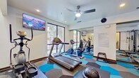 This fitness center is a thoughtfully designed workout facility that offers a comprehensive range of equipment to support diverse exercise routines. The space is finished with a striking hexagonal-patterned carpet in shades of blue, teal, and grey, lending a modern and energetic aesthetic throughout.

The cardio zone features treadmills, a stationary bike, and an elliptical machine, all positioned near large floor-to-ceiling windows that flood the room with natural light and provide views of the surrounding greenery. A ceiling fan overhead ensures comfortable air circulation during workouts.

The strength training area is well-stocked with a multi-functional cable machine, a full rack of dumbbells in graduated weights, a flat workout bench, and various functional training accessories including a medicine ball and step platforms. Wall-mounted mirrors along one side visually expand the space and allow users to monitor their form.

Multiple wall-mounted flat-screen TVs are strategically placed throughout the room, displaying nature-themed visuals for an immersive workout experience. A posted fitness policy guide and a wall clock keep users informed and on schedule. An adjacent restroom with a sink and stool provides added convenience. The overall ambiance is clean, bright, and functional, making this facility a well-rounded amenity for daily fitness needs.