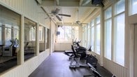 This fitness facility is housed in a charming, light-filled structure characterized by its distinctive architectural details. The space features white-painted tongue-and-groove wall paneling and exposed ceiling beams, lending it a classic yet functional character. Large multi-pane windows line the walls on multiple sides, flooding the interior with abundant natural light and offering views of the surrounding greenery, creating an inviting and energizing atmosphere for exercise.

The flooring consists of dark rubber gym matting, providing both durability and safety underfoot. Multiple ceiling fans are suspended from the beamed ceiling, ensuring consistent air circulation throughout the space. A wall-mounted television is positioned for easy viewing during workouts.

The room is equipped with a selection of cardio machines, including ellipticals, a treadmill, and a recumbent bike, all arranged in a row to maximize the available floor space. A mirrored wall section on one side enhances the sense of openness and allows users to monitor their form. The overall ambiance is bright, clean, and welcoming, making it a practical and pleasant environment for fitness activities.
