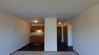 This single-floor space features an open-concept layout where the living area and kitchen are connected, separated by a partial dividing wall that adds structure without fully closing off either zone. The kitchen is equipped with warm-toned wooden cabinetry, both upper and lower units, providing ample storage. A sink is positioned along the counter, accompanied by under-cabinet lighting that illuminates the workspace. A small mat in front of the kitchen area adds a practical touch to the flooring, which contrasts with the carpeted living space. The living area is open and unobstructed, allowing for flexible furniture arrangement. A hallway visible beyond the dividing wall leads to additional areas of the unit, with two doors suggesting access to a bedroom and closet or bathroom. Flush-mount ceiling light fixtures provide consistent ambient lighting throughout. The textured white ceiling adds subtle visual interest overhead. The overall design is practical and straightforward, with a neutral color palette of whites and warm wood tones creating a simple, functional atmosphere suited to everyday living.