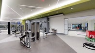 This contemporary fitness center offers a comprehensive workout environment designed to support a wide range of exercise routines. The space is defined by a clean, modern aesthetic, featuring a neutral gray and white color palette accented by bold lime-green architectural ceiling details that add energy and visual interest throughout.

The main workout floor is finished with durable, resilient rubber flooring in a speckled gray tone, providing both safety and comfort underfoot. A robust selection of strength training cable machines and multi-use equipment is arranged throughout the space, giving users access to a thorough resistance training experience. Mirrored wall panels line key areas, allowing users to monitor form during exercises while also amplifying the sense of openness.

Recessed ceiling lighting and sleek linear light fixtures provide bright, even illumination across the entire floor, ensuring excellent visibility at all times. A built-in water fountain station is conveniently positioned along the wall, offering easy hydration access.

A separate glass-walled studio or cardio room is visible toward the rear of the space, housing additional equipment such as stationary bikes or treadmills, and allowing for a distinct zone for different workout modalities. The transparent glass partition maintains a sense of visual connectivity while defining the separate functional areas. Overall, the fitness center presents a polished, professional environment well-suited for a focused and effective exercise experience.