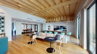 This clubhouse is a thoughtfully designed communal space that balances social gathering with focused work. The most striking architectural feature is the exposed wood beam ceiling structure that spans the main lounge area, adding warmth and visual depth to the open-plan layout. Light oak hardwood flooring runs throughout, reinforcing the space's cohesive, contemporary aesthetic.

The central lounge area is furnished with a neutral sectional sofa, modern accent chairs, and sleek black pedestal tables, creating an inviting environment for casual interaction. Teal upholstered seating lines a long communal bar-height table, offering an additional zone for collaborative work or socializing. A bold abstract artwork serves as a focal point along one wall, complemented by elevated built-in shelving stocked with books and décor.

A well-appointed kitchen features rich walnut cabinetry, a commercial-grade coffee station, stainless steel appliances, and a large island with a stone countertop, accented by a striking geometric tile backsplash. Glass-enclosed meeting rooms with black-framed sliding doors provide private workspaces while maintaining visual connectivity. A separate lounge nook with a deep channeled navy sofa overlooks what appears to be a pool area, adding a relaxed, resort-like quality to the overall ambiance.