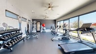 Located in Tucson, Arizona, this dedicated fitness center offers a thoughtfully designed exercise space that combines functionality with motivating aesthetics. The room is finished with durable speckled rubber flooring, providing a safe and supportive surface throughout the workout area.

A striking feature of the space is the dramatic dark accent wall adorned with large-scale athletic murals, creating an energizing atmosphere that encourages peak performance. Ceiling fans are installed overhead to promote air circulation, complementing the recessed lighting that keeps the space bright and well-lit.

The fitness center is comprehensively equipped for a full-body workout. Cardio equipment includes Spirit-brand treadmills and elliptical trainers, along with stationary and cycling bikes. Strength training is well-represented with a multi-functional cable and weight machine, a full rack of dumbbells with an adjustable bench, and a collection of stability and medicine balls for functional fitness exercises.

Large floor-to-ceiling windows line one wall, inviting abundant natural light and framing views of the surrounding Tucson landscape and distant mountain silhouettes. A set of glass double doors opens to an outdoor patio area, further connecting the interior to the scenic desert environment. The overall design is modern, clean, and purpose-built to support a wide range of fitness routines.