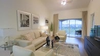 This single-floor residence offers a refined and welcoming atmosphere, thoughtfully designed with a neutral color palette and modern furnishings throughout. The living area serves as the centerpiece of the space, boasting impressively high ceilings that amplify the sense of openness and airiness. Large sliding glass doors flood the room with abundant natural light and provide seamless access to a private balcony, where sweeping views of verdant greenery create a tranquil outdoor retreat. The living room is elegantly furnished with cream-toned sofas and armchairs arranged around a sleek glass coffee table, complemented by decorative accents including sculptural pieces and potted plants that add warmth and character. Black-and-white framed artwork adorns the walls, lending a sophisticated, curated aesthetic to the space. A ceiling fan with a polished fixture enhances comfort while maintaining the room's clean, contemporary style. The overall ambiance is one of calm sophistication — bright, airy, and thoughtfully appointed, making it an ideal environment for both relaxed everyday living and stylish entertaining.