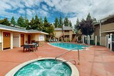 Located at 591 Peabody Rd in Vacaville, California, this single-story outdoor amenity facility serves as a communal recreational hub. The space is centered around a generously sized swimming pool with clearly marked depth indicators and a surrounding terracotta-toned concrete deck that provides ample room for lounging. Adjacent to the pool is a bubbling spa/hot tub, perfect for relaxation. A set of teal-colored chaise lounges and outdoor dining sets are arranged throughout the deck area, offering comfortable spaces for sunbathing and socializing.

The building itself features a Craftsman-inspired architectural design, with wood panel siding, stone accents, pitched gabled rooflines, and warm-toned painted doors. Inside, the facility includes two restrooms — designated for separate use — as well as a utility room. Colorful mural artwork decorates the exterior facade, adding a vibrant, welcoming touch to the space.

A covered pavilion with a pergola-style roof and built-in BBQ grill provides a shaded outdoor kitchen and dining area, ideal for gatherings. The surrounding landscape is lined with mature trees, including ornamental and deciduous varieties, which frame the space with natural greenery. The pool area is enclosed by a fence, ensuring privacy and security for users of this well-appointed community amenity space.