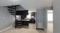 This two-story residence at The Fields offers a well-designed layout that balances functionality with contemporary style. The main level showcases an open-concept kitchen equipped with dark espresso cabinetry, black appliances including a range and refrigerator, and a kitchen island that provides additional workspace and casual dining potential. A window above the sink allows natural light to brighten the cooking area, while wood-look flooring runs throughout the kitchen, creating a warm, cohesive aesthetic.

The transition between the kitchen and adjacent living areas is seamless, with a combination of hard flooring and carpet defining distinct zones within the space. Neutral gray walls provide a versatile backdrop throughout, complementing the dark tones of the cabinetry and fixtures.

One of the most distinctive architectural features is the spiral staircase, finished in dark metal with wood-toned steps, which connects the main level to the upper floor. Its compact, elegant design makes an immediate visual statement while efficiently utilizing vertical space. The textured ceiling adds depth overhead, and thoughtful details such as a wall-mounted thermostat and floor-level vents reflect the home's practical, move-in-ready condition. Overall, the space presents a modern, coordinated interior suited for comfortable everyday living.